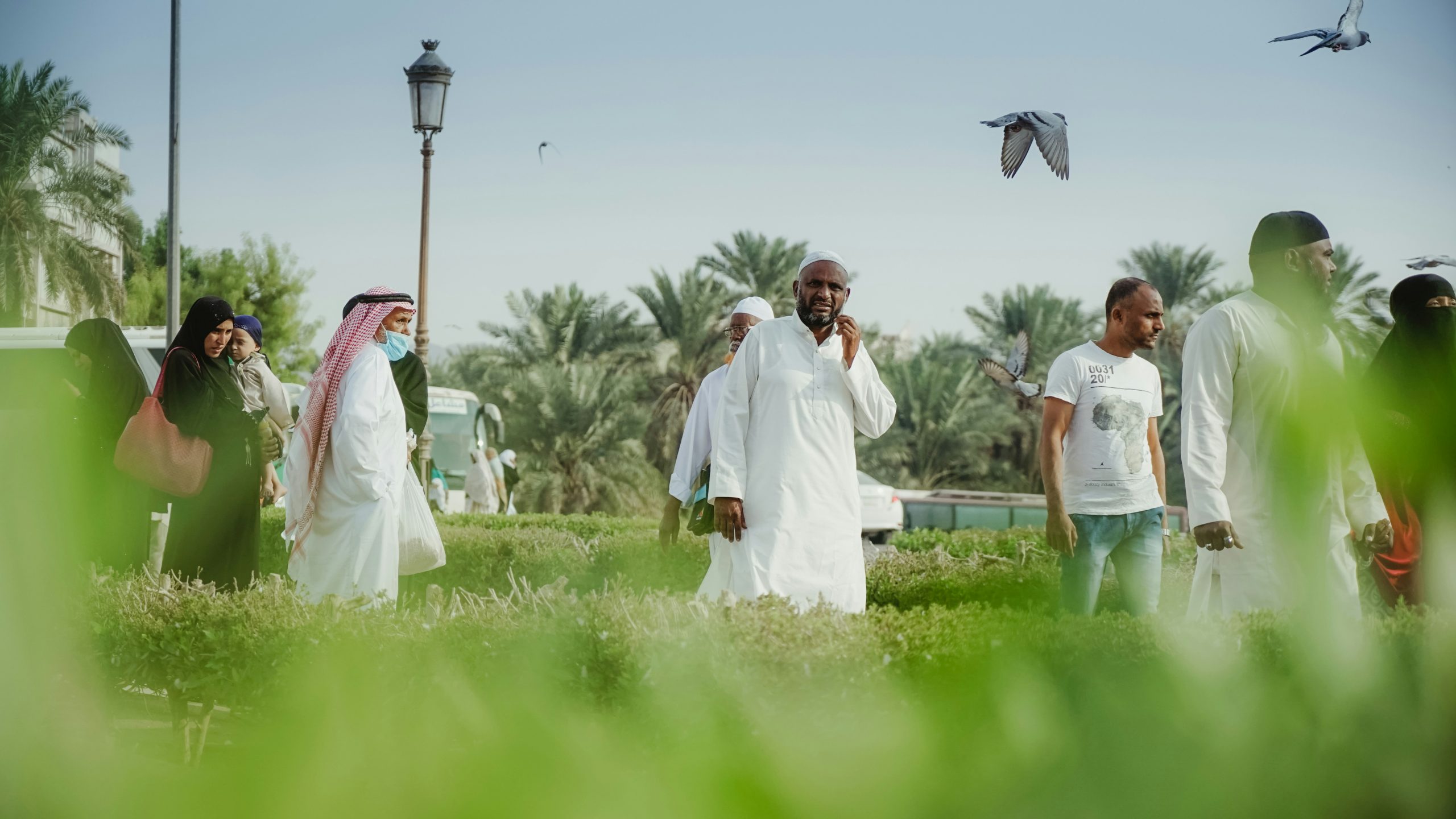 People standing. Some men are wearing thawbs (white, long sleeve, ankle length robe). One is wearing a keffiyeh (red and white headscarf). The women are wearing long sleeve black robes, two are wearing hijab, another is wearing a niqab. There is one man wearing a tshirt and jeans.