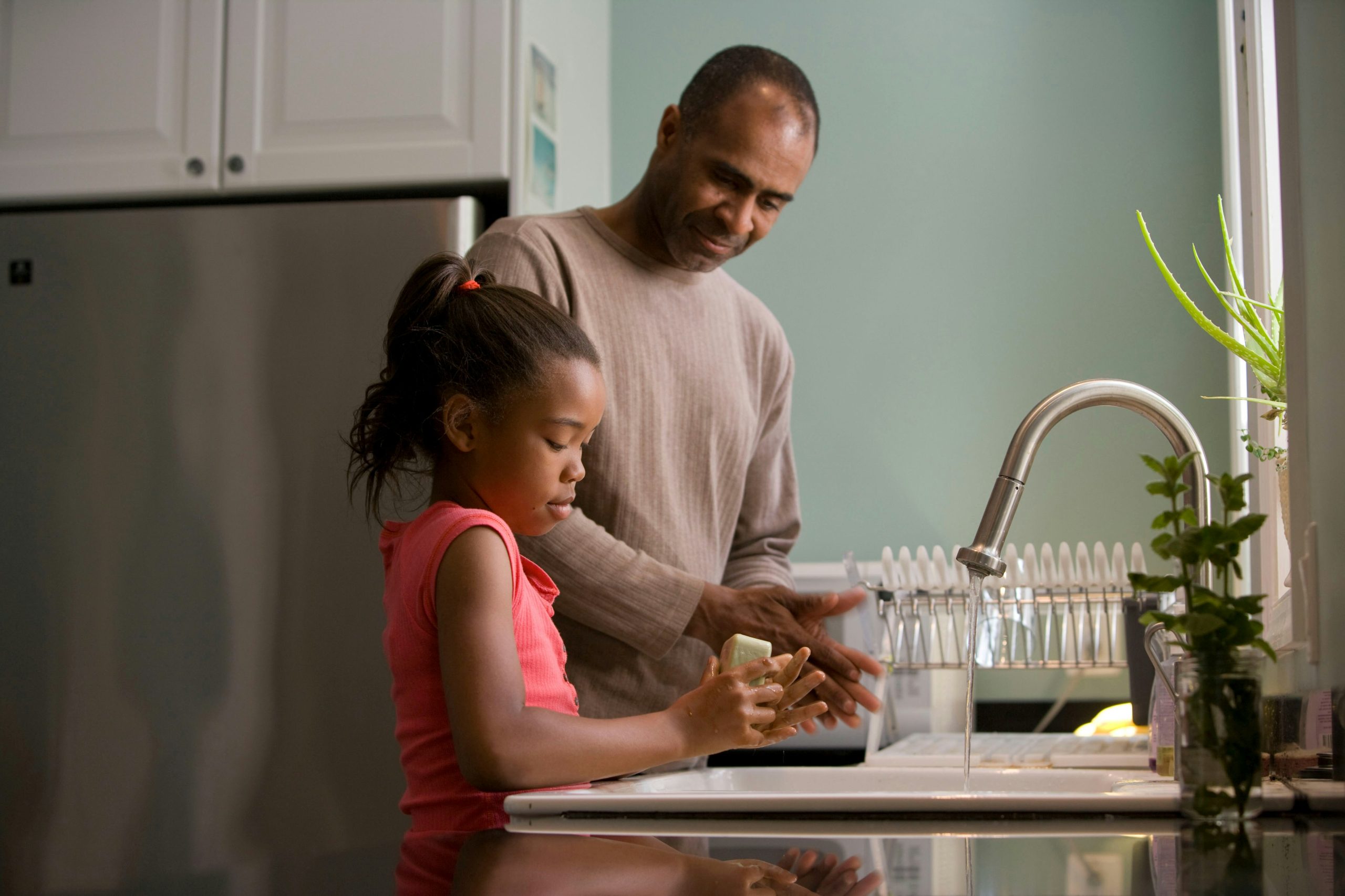 A father watching his young daughter washing her hands in the kitchen sink with bar soap