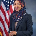 Headshot of a hijabi woman (Ilhan Omar) in a suit smiling in front of an American flag, typical of congressperson headshots.