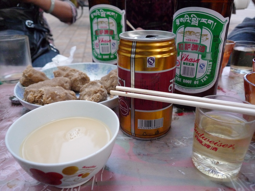 Taken from the perspective of someone sitting at a table: Tibetan beer, chopsticks resting on a cup, a latte-colored drink, and food on a plate