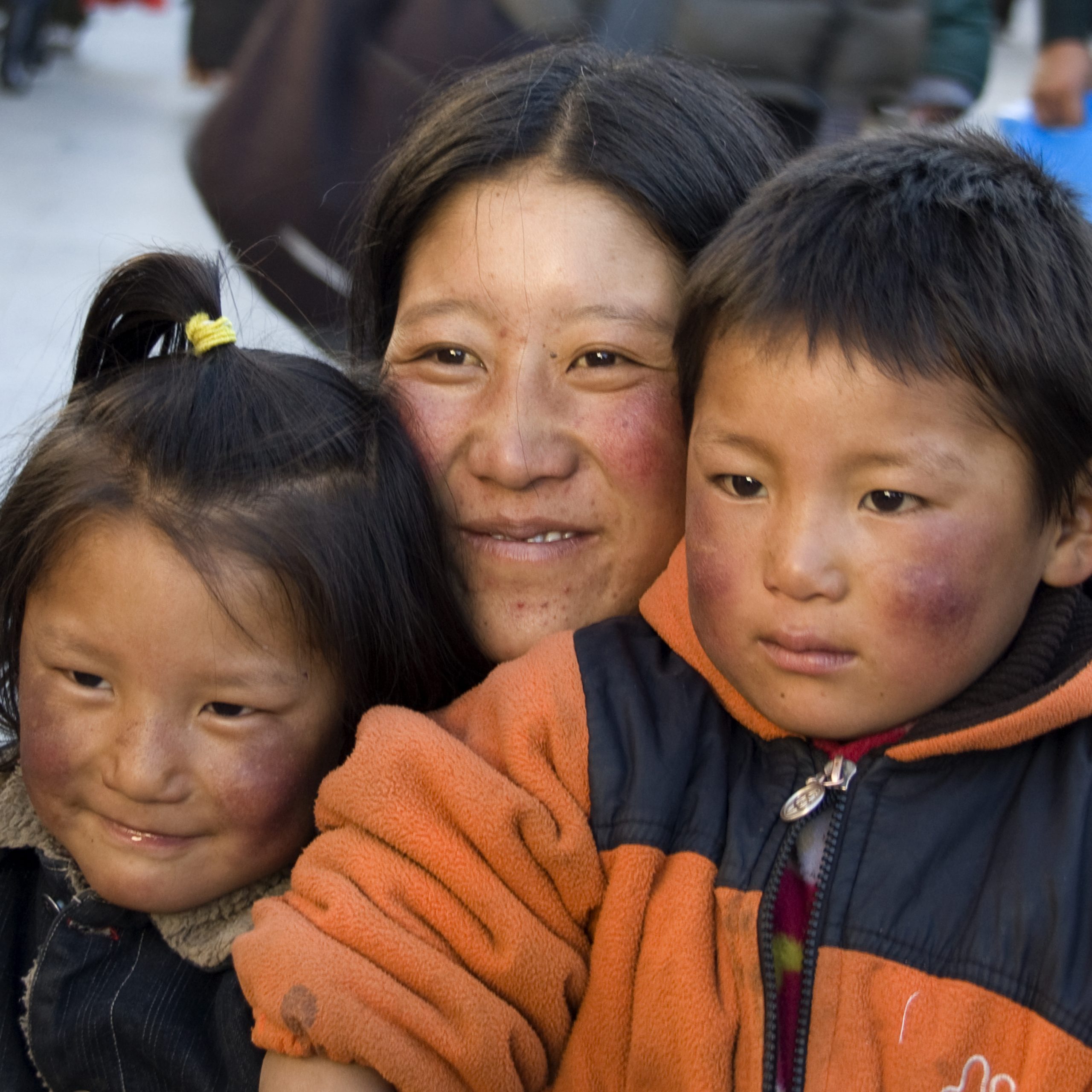 Tibetan mother crouching down to stand with her girl and boy children. They all have red cheeks. The mother and the girl are smiling
