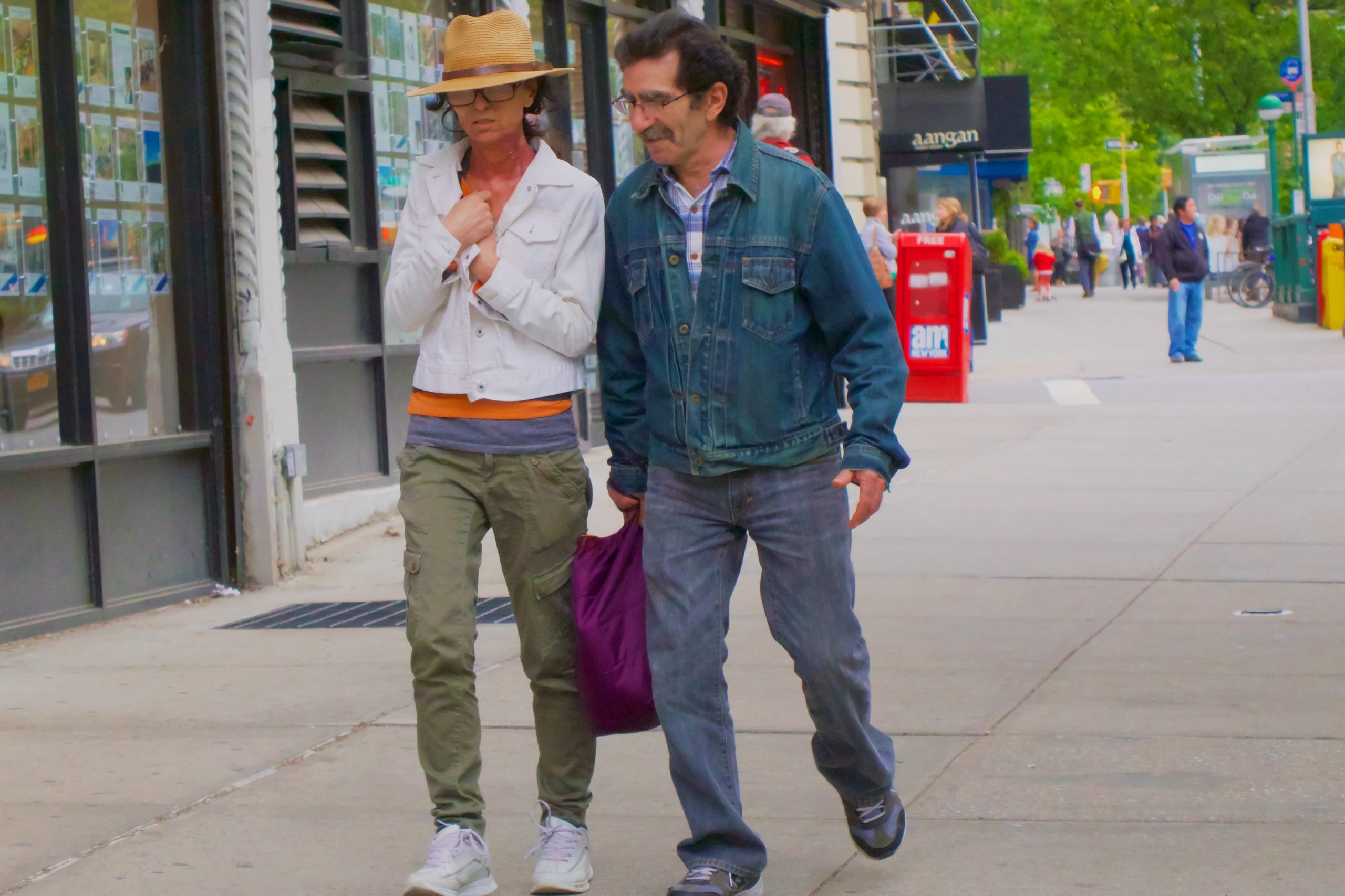 A man and woman are walking down a city street. The woman appears to be wearing glasses and a hat. She is dressed in an orange-gray colour pattern t-shirt and a white jean jacket. She's also wearing green jean pants and white sneakers. The man also appears to wear eyeglasses. He's wearing a pattern shirt and a blue jacket. He's also wearing grey jean pants and grey sneakers.