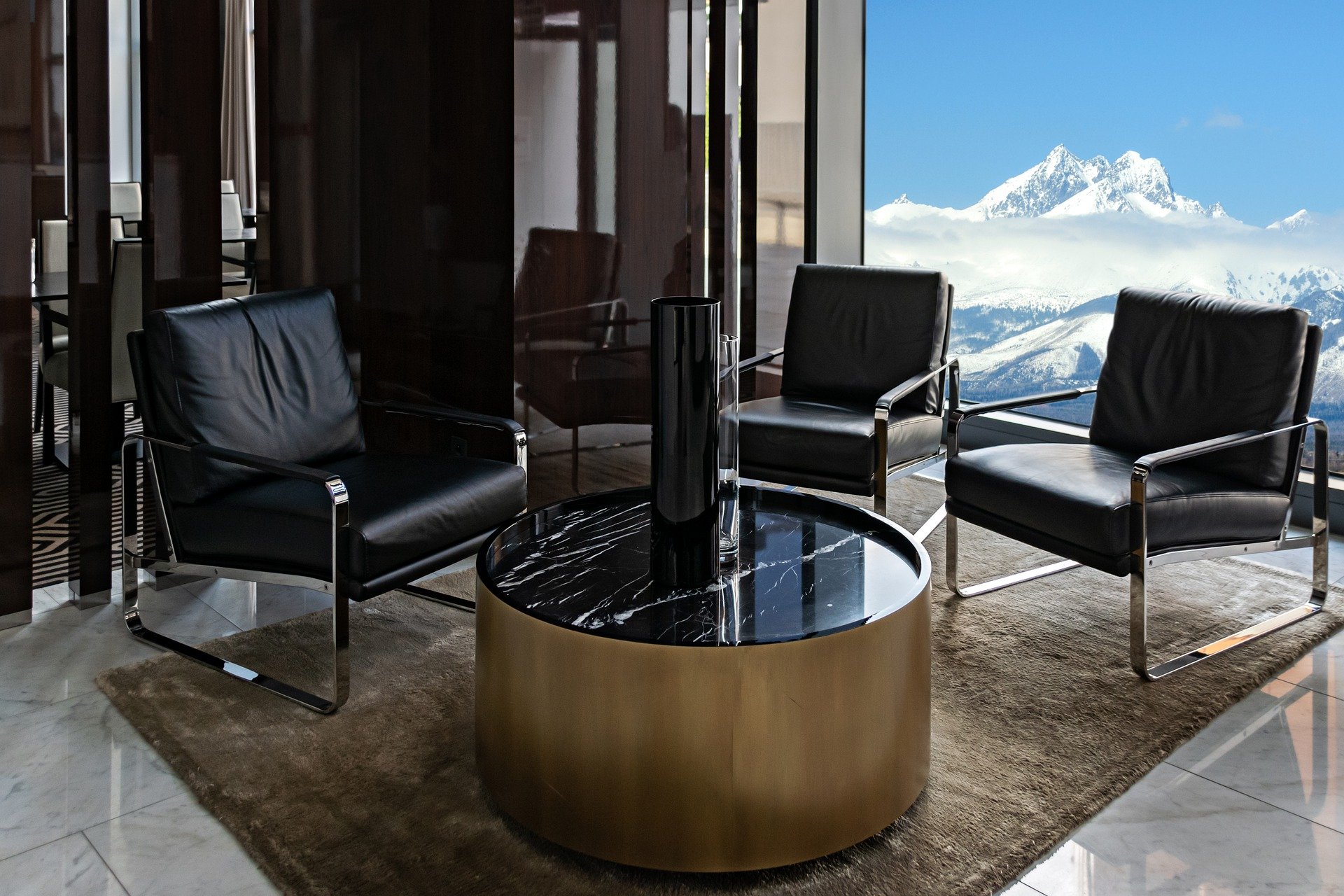 The image illustrates a modern living room area with three black leather armchairs and a round, black marble-topped coffee table, overlooking a panoramic view of snow-capped mountains through a large window.