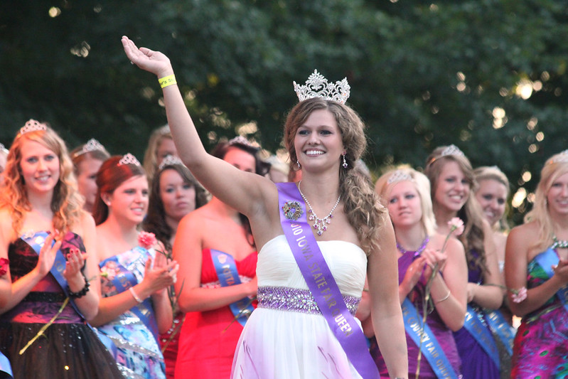 woman wearing an iowa state fair queen sash smiling and waving standing in front of other female contestants