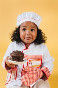 a little girl with dark curly hair wearing a chef outfit and holding a cupcake in one hand and mitten in other hand sitting on a stool.