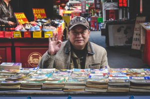 Man in Brown Coat in Front of Books, selling books